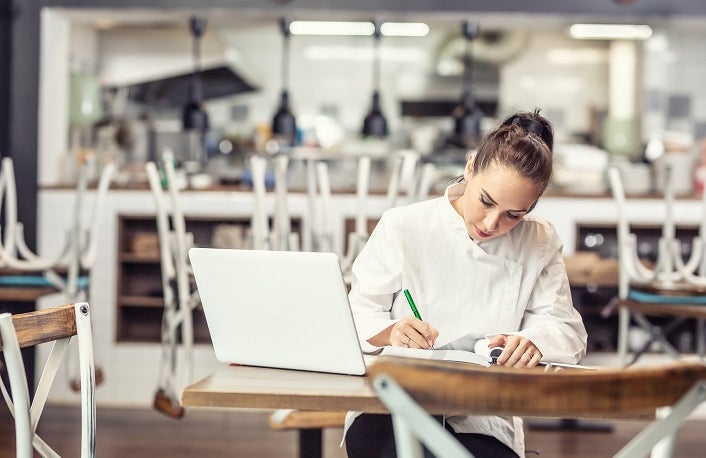 Female restaurant chef sits at the table after hours doing her bookkeeping in front of an open laptop.