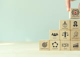 A single hand placing a small wooden block against a tower of blocks. Each block has a separate core value printed on them.