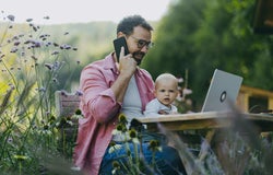 Man working on a laptop and talking on a phone while holding a baby