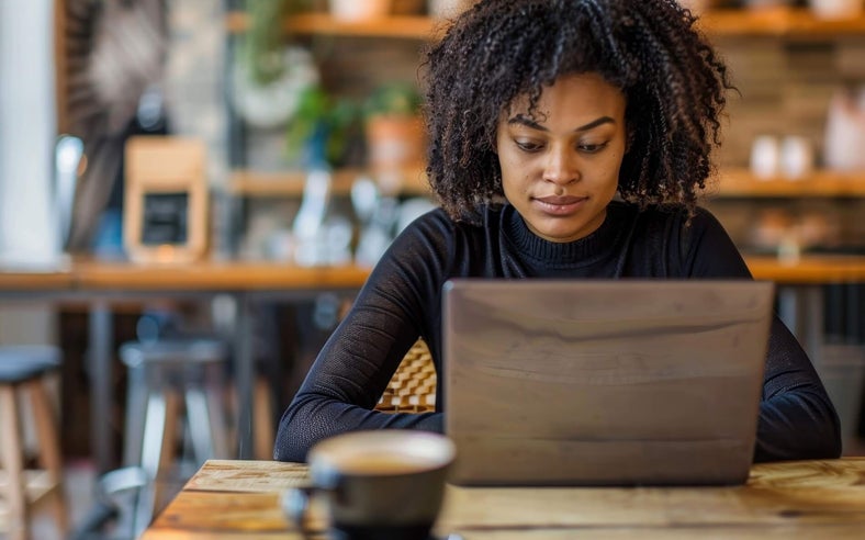 A woman with a black sweater sitting at wooden cafe table with a brown laptop.