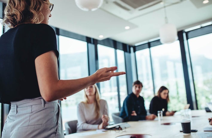 A back shot of a blonde woman in a black top and grey trousers addressing team members in a meeting room.